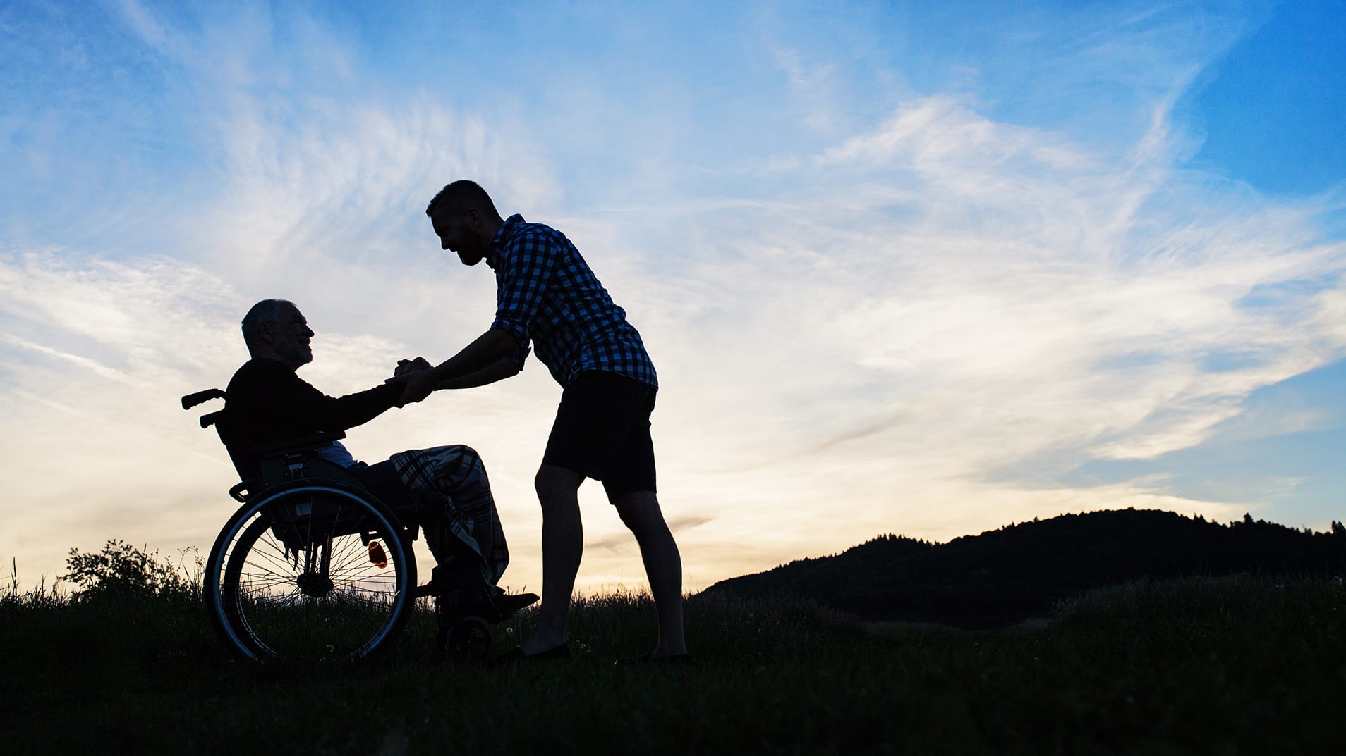 A silhouette of adult son looking at his father in wheelchair in nature at sunset.